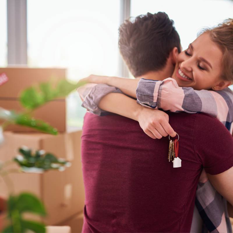 Pareja joven vestida casual en la que ella lo abraza a él mostrándose contenta mientras sostiene un juego de llaves en la mano y varias cajas de mudanza al fondo aún sin abrir, como si acabasen de alquilar un piso para larga temporada en Fuengirola