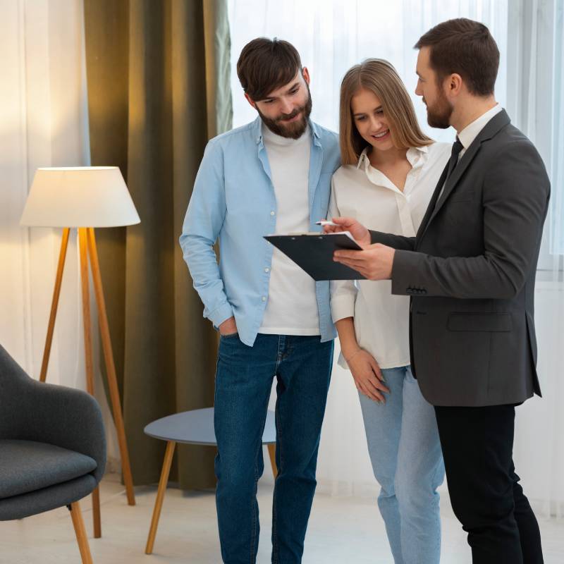 Agente inmobiliario masculino vistiendo traje, con barba cuidada y bien peinado, con un portfolio y bolígrafo en la otra mano explicándole a una pareja joven las condiciones de venta de una casa o piso en Fuengirola, en cuyo salón, amueblado con estilo moderno, se encuentran de pie los tres.